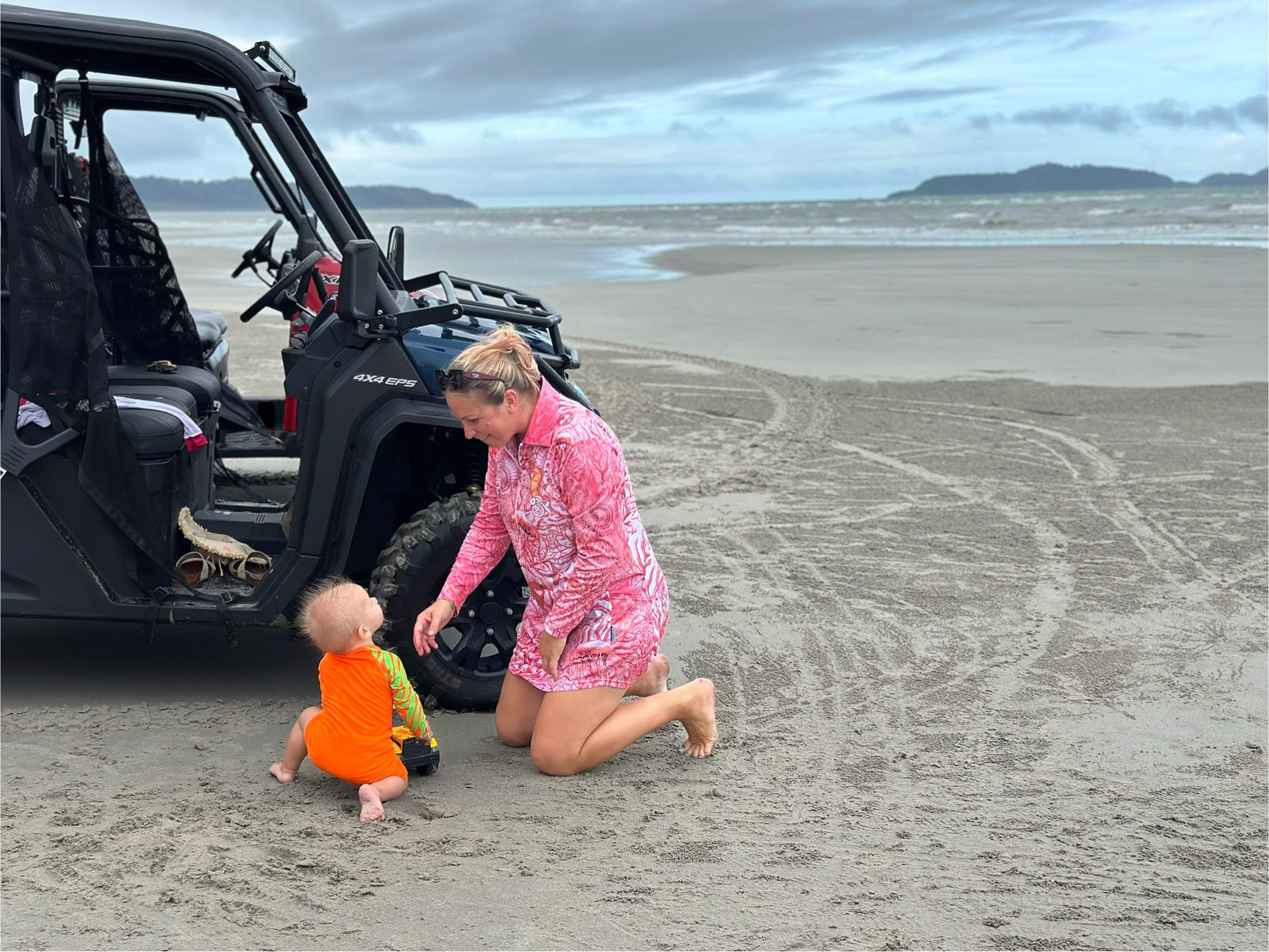Woman wearing pink fishing dress with nursing zip, playing with toddler by ATV on a sandy beach in Far North Queensland, Australia