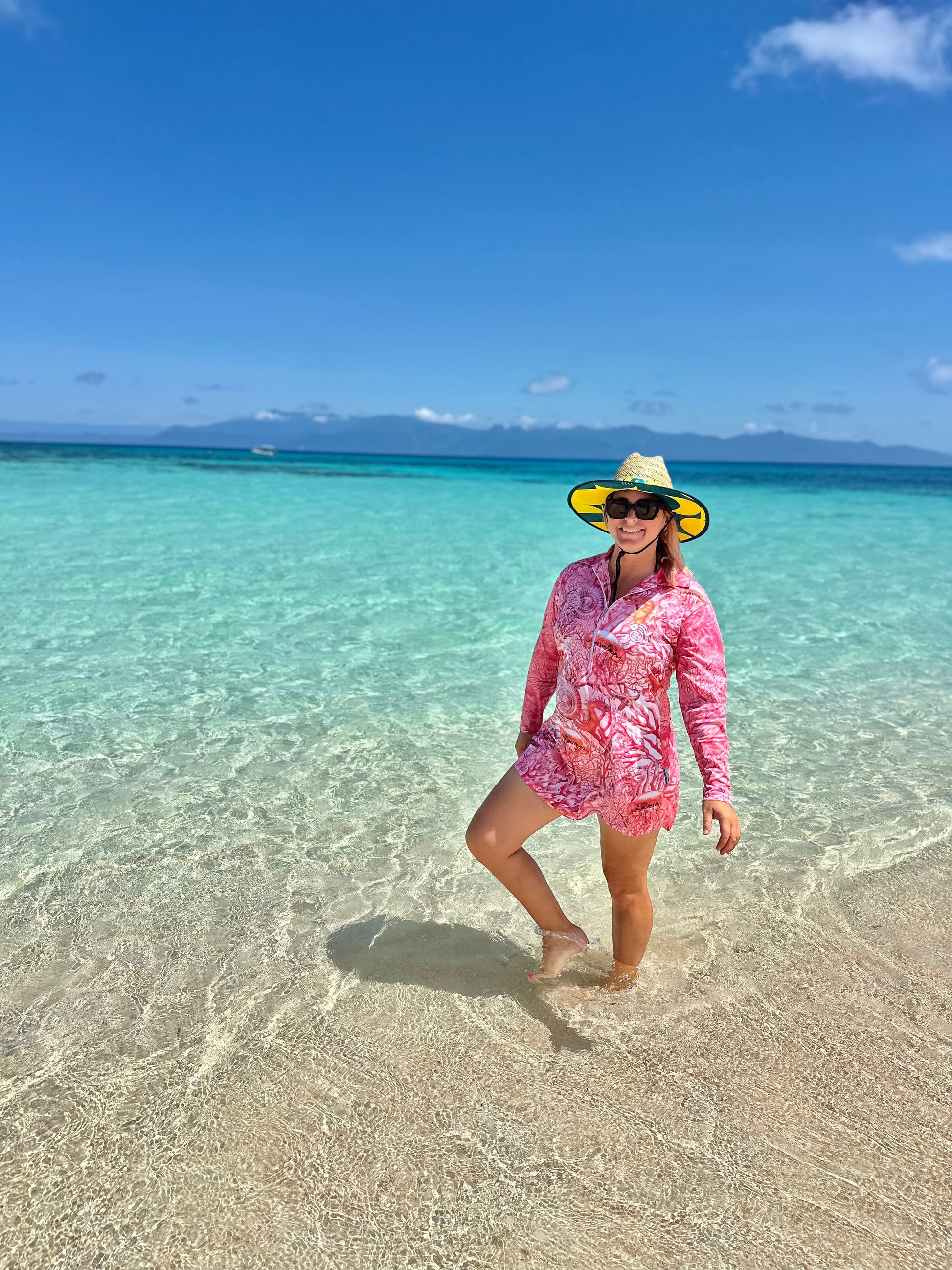 Woman in a 'Fish Feeder Wear' pink fishing dress with nursing zip and hat standing in clear turquoise water with a blue sky on the Great Barrier Reef in Australia