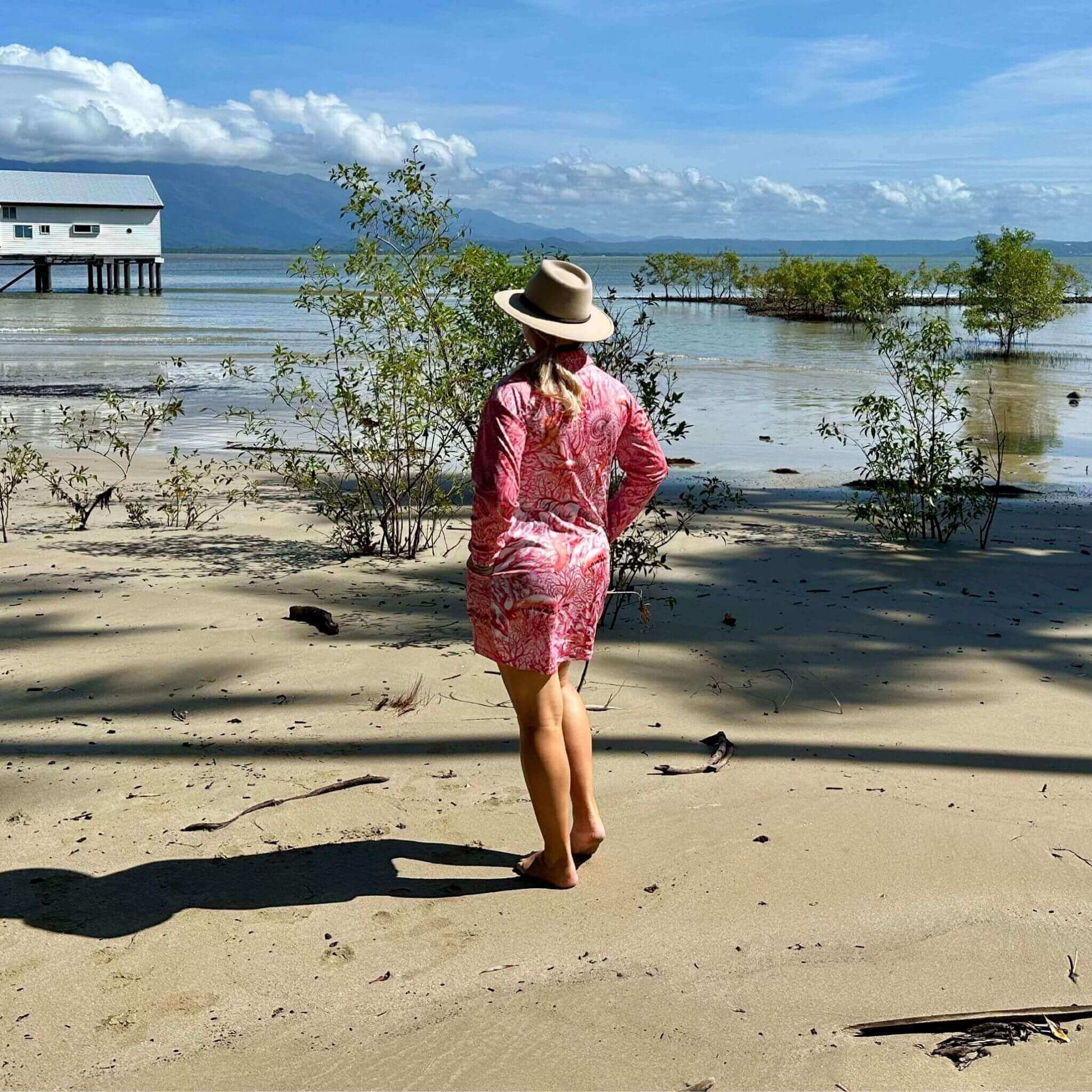 Woman in a 'Fish Feeder Wear' pink fishing dress and hat standing on a sandy beach with Sugar Wharf, trees and water in the background in Port Douglas, Australia.