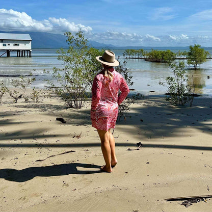 Woman in a 'Fish Feeder Wear' pink fishing dress and hat standing on a sandy beach with Sugar Wharf, trees and water in the background in Port Douglas, Australia.