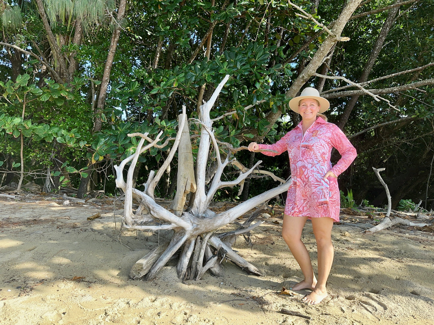 Woman in a 'Fish Feeder Wear' pink fishing dress with nursing zip standing next to a large piece of driftwood on a beach with trees in the background in Far North Queensland, Australia