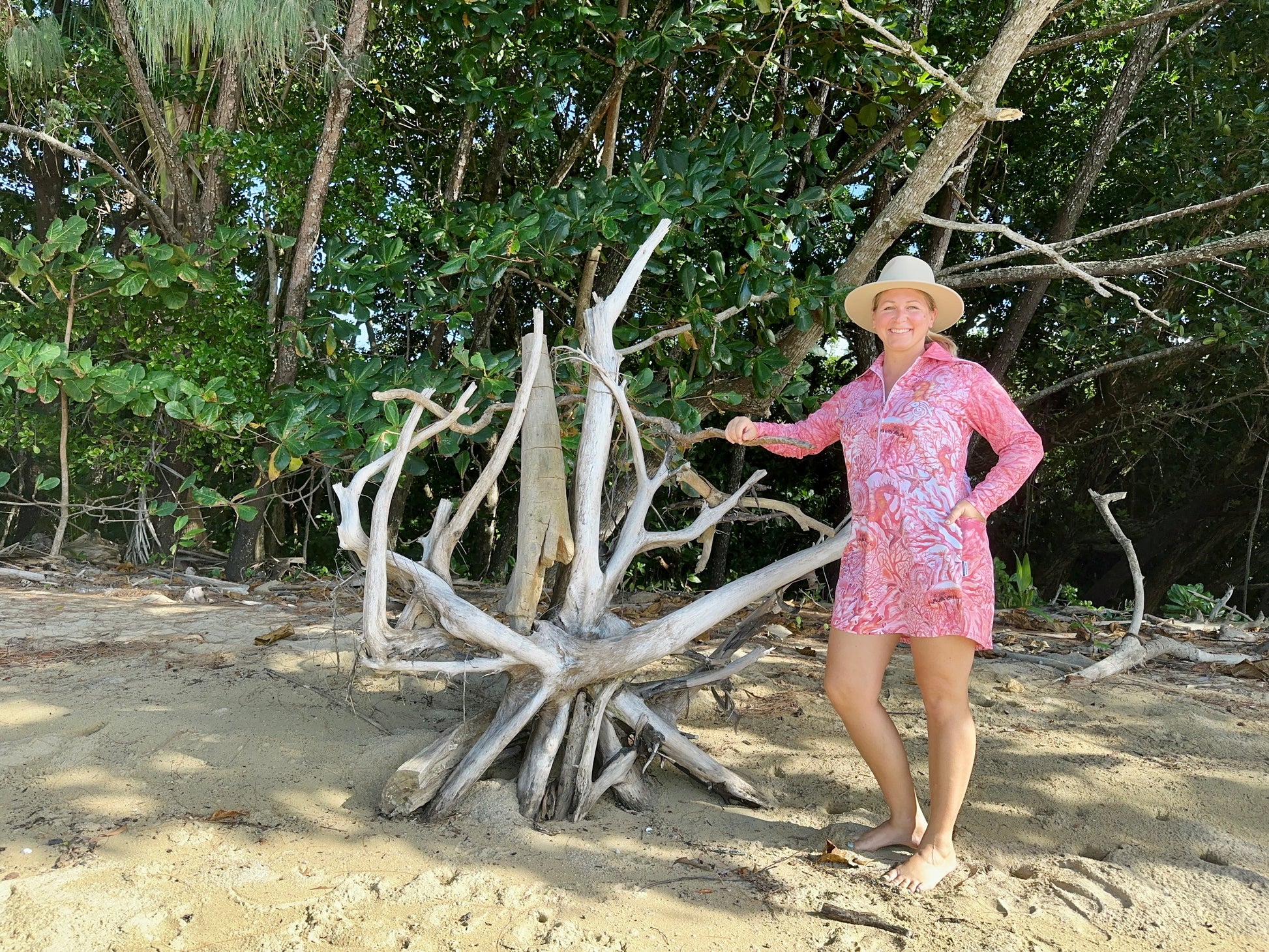 Woman in a 'Fish Feeder Wear' pink fishing dress with nursing zip standing next to a large piece of driftwood on a beach with trees in the background in Far North Queensland, Australia