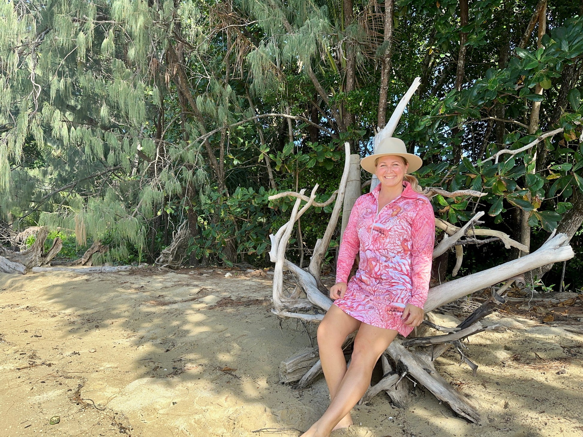 Lady in a 'Fish Feeder Wear' pink fishing dress with nursing zip sitting on driftwood with greenery in the background in Far North Queensland, Australia