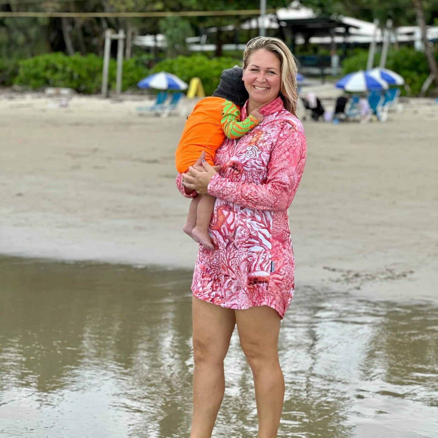 Woman in 'Fish Feeder Wear' pink fishing dress with nursing zip holding a baby on a beach in Port Douglas, Australia